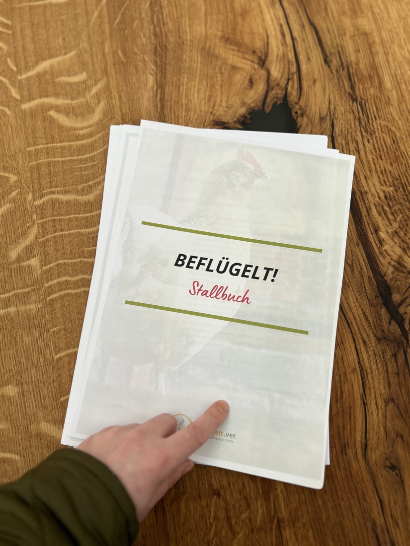 Das ausgedruckte beflügelt Stallbuch liegt auf einem Holztisch, Dr. Eva-Maria Casteel zeigt auf das Deckblatt